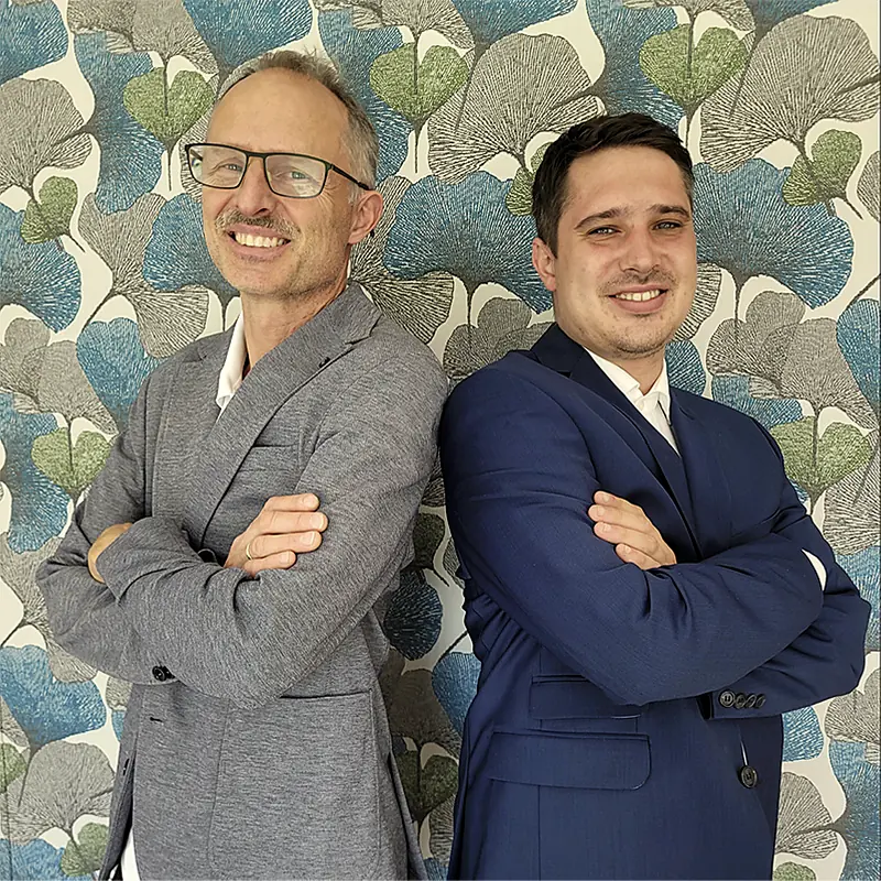 Manfred und Florian Sebald stehen Rücken an Rücken vor einer gemusterten Tapete