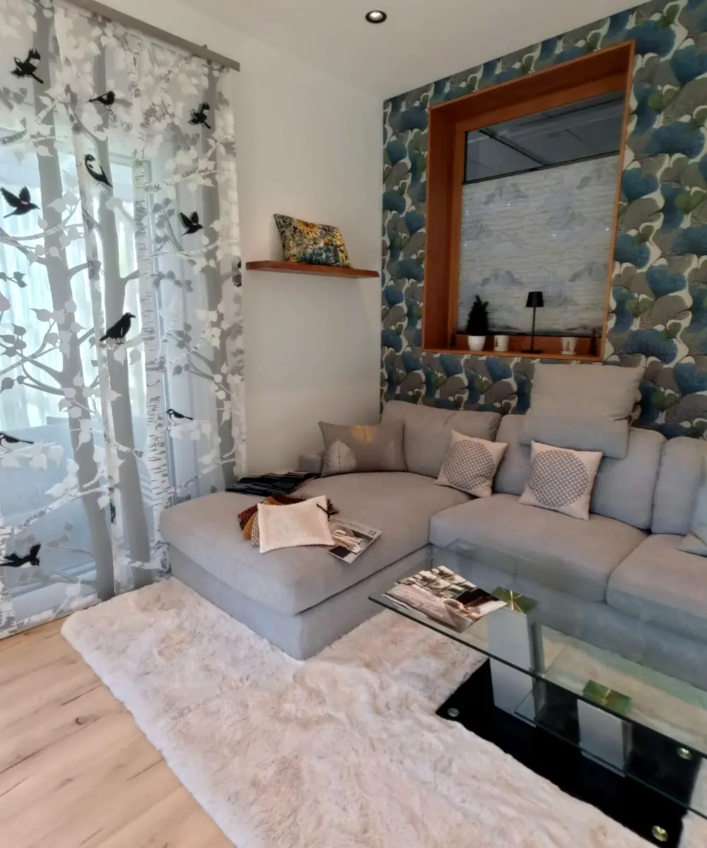 Sitzecke im Showroom: graues Sofa, gemusterte Tapete, Flächenvorhang mit Birkenmotiv