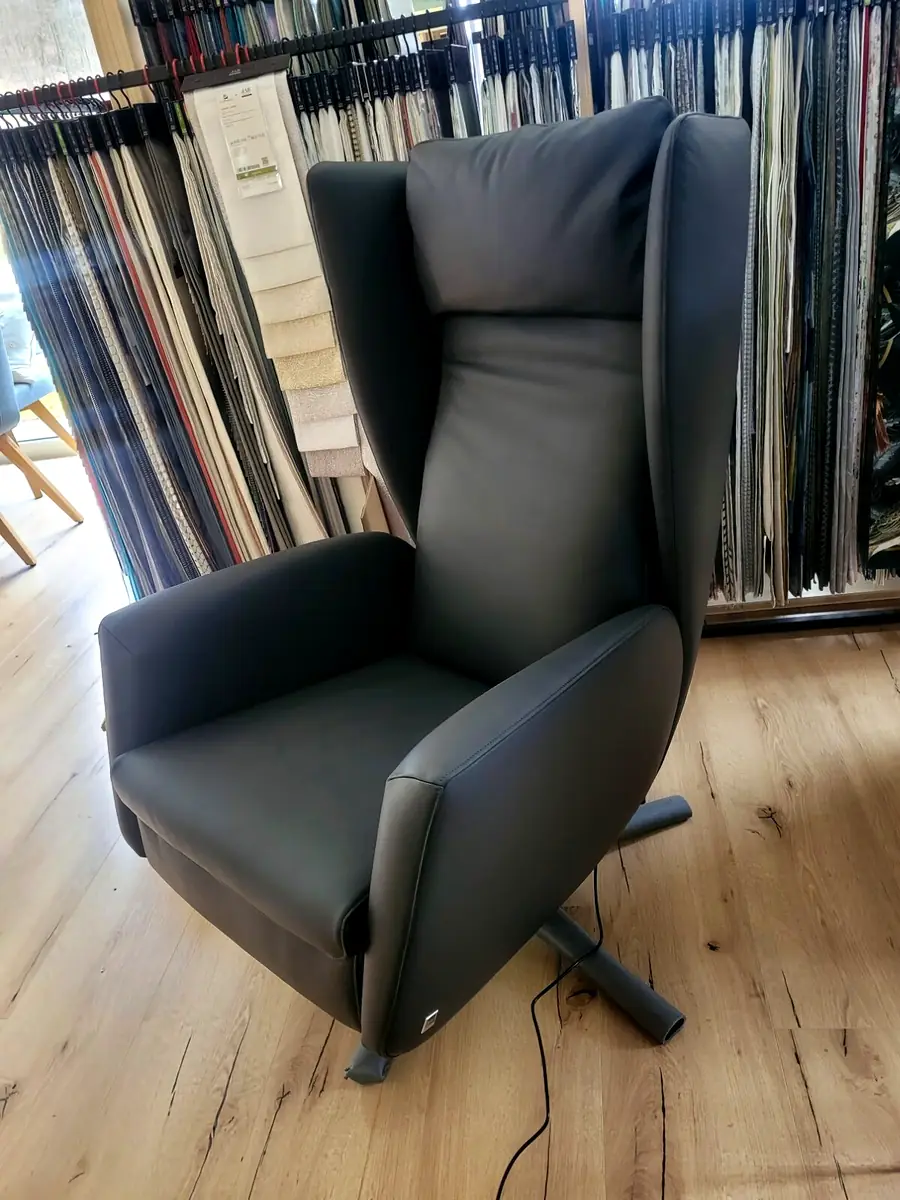 Hochlehner-Sessel mit dunklem Lederbezug vor einer Wand voller Stoffmuster