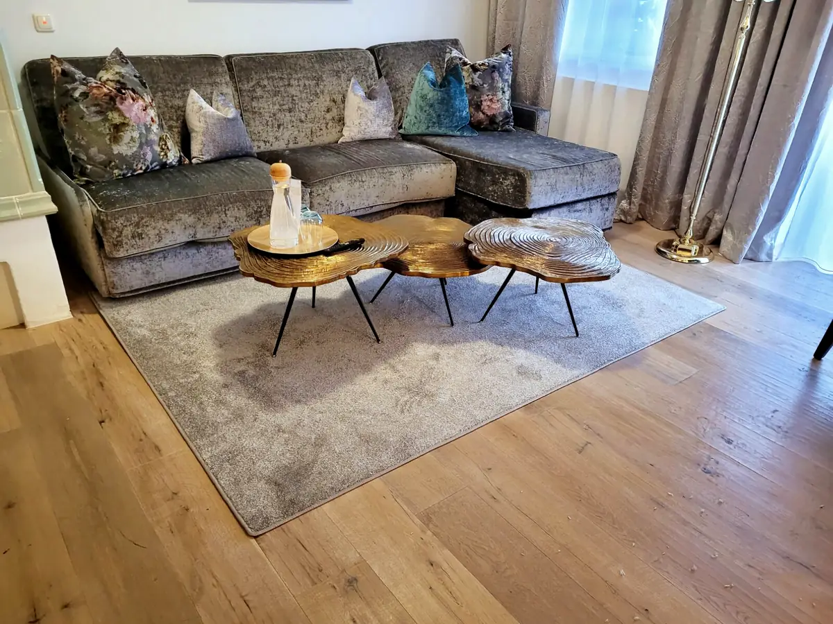 Ecksofa mit schimmerndem Samtbezug und gemusterten Kissen in einem Wohnzimmer, davor ein Teppich und Holztische